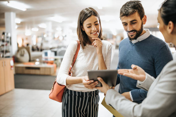 Young couple buying new furniture and looking for choices on digital tablet with help of sales person at the store.